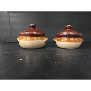 Op de afbeelding: Twee kleine keramische potten met deksel. De potten hebben een lichtbeige basis, een oranjebruine band en een donkerbruin deksel met een kleine knop. De potten hebben kleine handvatten aan weerszijden. De achtergrond is donker.