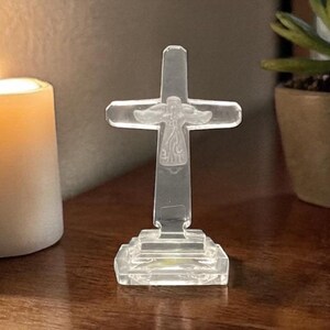 May include: A clear, transparent cross figurine with an etched figure in the center. The cross is mounted on a rectangular base. The background includes a lit candle and a potted plant.