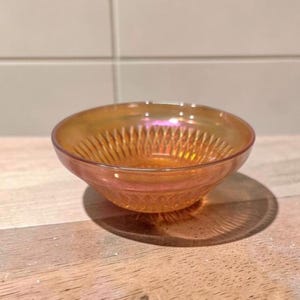 May include: An iridescent orange glass bowl with a decorative pattern. The bowl has a rounded shape and sits on a wooden surface. The bowl's interior features a detailed, textured design.