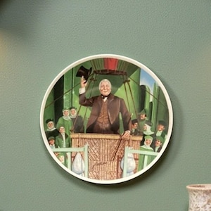 May include: A decorative plate showing a scene from a classic story. The plate depicts a man in a hot air balloon basket waving to a crowd. The plate has a white border and is mounted on a green wall.