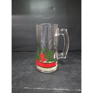 May include: A clear glass beer mug with a handle, decorated with a red ribbon and green holly leaves and red berries. The mug is cylindrical and has a wide base. The design is near the bottom of the mug.