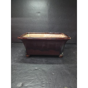 May include: A rectangular, dark brown ceramic planter with a glossy finish. The planter has a slightly flared shape and sits on four small, rounded feet. The interior is a lighter tan colour.