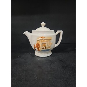 Könnte beinhalten: Weiße Keramik-Teekanne mit Deckel und Henkel. Die Teekanne zeigt eine dekorative Szene eines Kamins und eines Spinnrads in Orange-, Braun- und Blautönen. Die Teekanne ist im Vintage-Stil gehalten und steht auf einem Sockel.