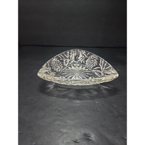 May include: Clear, triangular-shaped glass ashtray with intricate floral and swirl patterns. The ashtray has a central starburst design and a textured rim. The glass is transparent, allowing light to pass through, and sits on a dark surface.