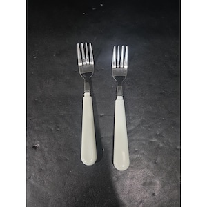 May include: Two stainless steel forks with ivory-colored handles are displayed against a dark background. The forks have four tines each and a classic design, suitable for everyday dining. The handles are smooth and tapered.