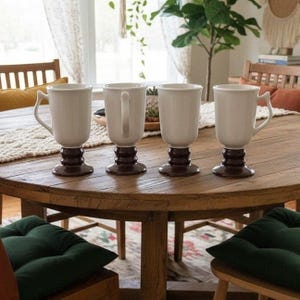 Puede incluir: Cuatro tazas de cerámica con un cuerpo superior blanco brillante y una base de pedestal escalonada de color marrón oscuro. Cada taza presenta un asa curva en el lateral. Las tazas están dispuestas en fila sobre una superficie de madera.