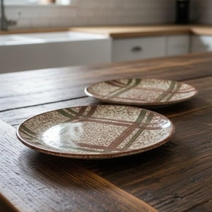 May include: Two round ceramic plates featuring a speckled cream base with a brown and muted green plaid pattern. The plates have a smooth, glossy finish and a low profile, showcasing a consistent design across both pieces of dinnerware.