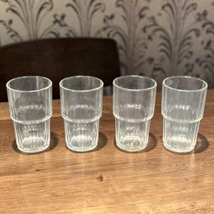 May include: Four clear, stackable drinking glasses with a ribbed design. The glasses are made of transparent glass and sit on a wooden surface. The glasses are empty and ready to be filled with a beverage.