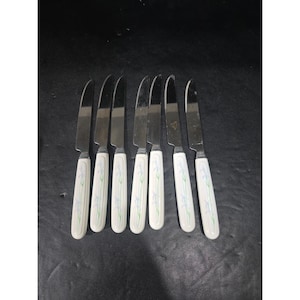 May include: A collection of seven butter knives. Each knife features a stainless steel blade and a white handle adorned with a floral design in blue and green. The knives are displayed on a dark surface.