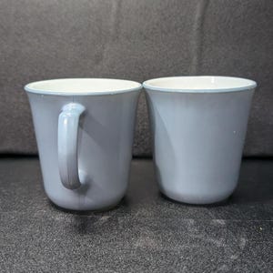 Two light blue coffee mugs placed next to each other, with the left mug showing its handle profile and the right mug facing forward.