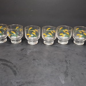 May include: Six clear glass shot glasses with a gold rim and a design of green wheat stalks and yellow wheat heads.