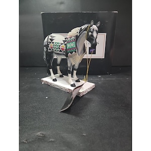 Op de afbeelding: Een decoratief paardenbeeldje met een zwart-witte vacht, versierd met een kleurrijk patroon. Het paard staat op een witte basis met een decoratieve rand. Een goudkleurig touwtje is aan de kop van het paard bevestigd.