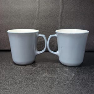 Two light blue coffee mugs standing side by side, both angled slightly with handles visible on the outer sides.