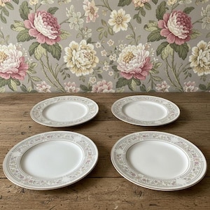 May include: A set of four round white ceramic plates, each featuring a delicate border pattern of small pink and blue flowers with green leaves and thin grey lines. The plates have a smooth, plain white center and a slightly raised rim edge.