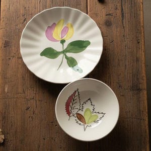 May include: A white ceramic plate with a scalloped edge and a painted yellow and pink flower with green leaves, paired with a smaller white ceramic bowl featuring a black line drawing of leaves with red and green accents on the interior surface.