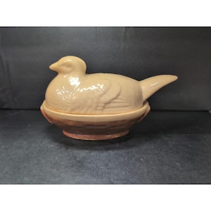 以下が含まれることがあります： 明るいタン色の蓋付きのセラミック製の鳥型容器。鳥は丸い茶色の台座に止まっています。この容器は、装飾または保管目的で使用される可能性があります。