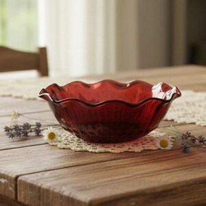 May include: A deep red glass bowl featuring a scalloped, wavy rim and a ribbed exterior texture. The bowl is translucent, allowing light to pass through its curved walls, and it sits centered on a circular, cream-colored crocheted lace doily.