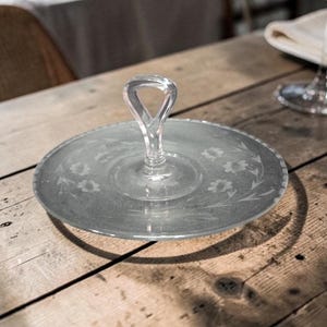 Op de afbeelding: Een heldere glazen serveerschaal met een handvat. De schaal heeft een cirkelvorm met geëtste bloemmotieven rond de rand en op het oppervlak. De handgreep is lusvormig. De schaal staat op een houten oppervlak.