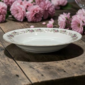 May include: White ceramic serving bowl with a floral pattern of pink and green flowers and leaves around the rim. The bowl has a gold trim and sits on a wooden surface.