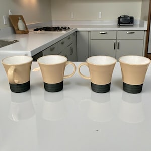 May include: Four beige ceramic mugs with black bases and white interiors are arranged on a white surface. Each mug has a curved handle and a slightly flared shape. The mugs are evenly spaced and reflect on the glossy surface.