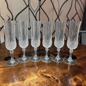 May include: Six clear crystal champagne flutes. Each flute features a diamond-cut pattern on the bowl and a slender stem. The glasses are arranged on a dark wooden surface, showcasing their elegant design. The flutes are ideal for celebratory occasions.