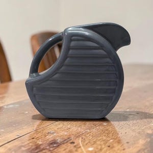 May include: A gray ceramic pitcher with a curved handle and a spout. The pitcher has horizontal ridges on the body and sits on a wooden surface. The background includes a wooden chair.
