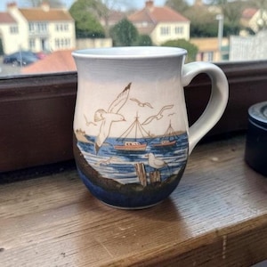 Peut inclure: Mug en céramique avec un motif côtier. Le mug présente un ciel bleu et blanc, une mer bleue, des mouettes et des bateaux. La poignée est blanche et le mug est posé sur une surface en bois.