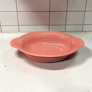 May include: A coral-colored ceramic serving bowl with two handles. The bowl has a smooth, glossy finish and a shallow, oval shape. The bowl is sitting on a white surface.