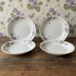 May include: A set of four white ceramic bowls featuring a delicate floral pattern of small purple violets and green leaves arranged along the curved rims. The bowls have a smooth, glossy finish and a shallow, rounded shape for serving food.