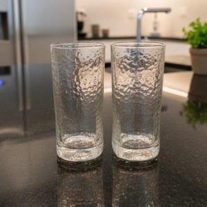 Op de afbeelding: Twee heldere, getextureerde drinkglazen. De glazen zijn cilindrisch met een licht getextureerd oppervlak. Ze staan op een donker aanrecht, met een keuken en een plant op de achtergrond.