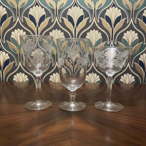 May include: Three clear glass vessels with etched floral patterns. The set includes a stemmed wine glass, a tall water goblet, and a wide-rimmed coupe glass, each featuring delicate white flower engravings around the upper portion of the bowls.