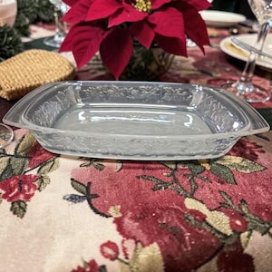 May include: A clear, rectangular glass serving dish with a floral pattern around the edges. The dish is set on a floral tablecloth, with a poinsettia and other table settings in the background. The dish is approximately 10 inches long and 6 inches wide.