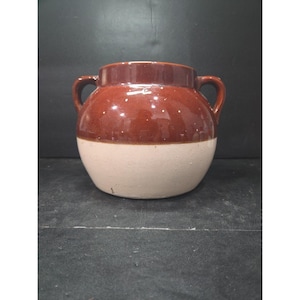 Puede incluir: Un tarro de cerámica de dos tonos, marrón en la parte superior y crema en la inferior. Tiene dos asas pequeñas y una abertura ancha. La sección marrón tiene pequeñas motas blancas. Probablemente para almacenamiento o decoración.