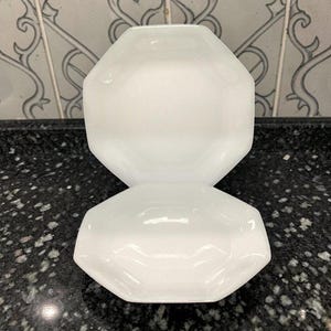May include: Two white, octagonal-shaped glass bowls stacked on top of each other. The bowls have a glossy finish and are sitting on a black speckled surface. The background features a decorative tile pattern.