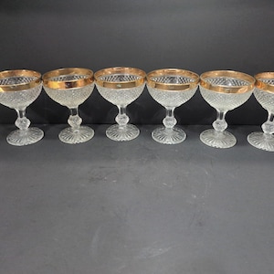 May include: Set of six clear glass champagne coupes with gold rims. The glasses have a diamond-shaped pattern and are on a black surface.