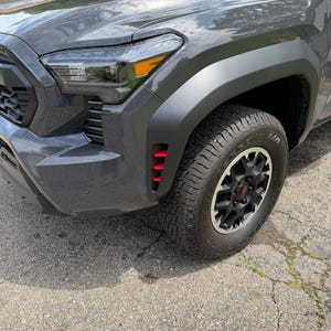 May include: A dark grey pickup truck front fender and wheel assembly. The vehicle features a matte black wheel arch, a black alloy wheel with a red centre cap, and a vertical vent with three bright red horizontal slats integrated into the fender.