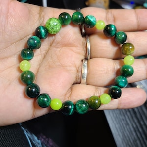 Op de afbeelding: Een armband met groene en limoenkleurige kralen. De armband heeft tijgeroogkralen en een glinsterende groene kraal. De armband is geregen op een elastische band.