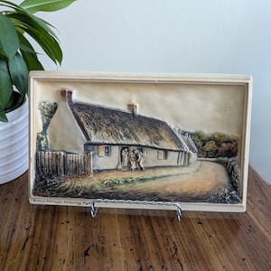 May include: A rectangular, cream-colored relief plaque depicting a thatched-roof cottage scene. The artwork features a winding road, a wooden fence, and figures near the cottage entrance. The text "Burns' Cottage, Alloway Ayr" is visible.