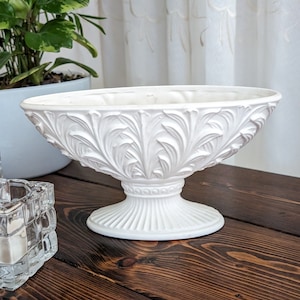 May include: White ceramic bowl with an ornate leaf design. The bowl is on a pedestal base and has a smooth, glossy finish.