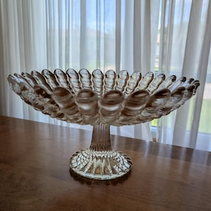 May include: A clear glass pedestal bowl featuring a wide, shallow basin with a scalloped, ribbed edge. The bowl stands on a thick, vertically fluted stem that connects to a circular, textured base, creating a structured and elegant display piece.