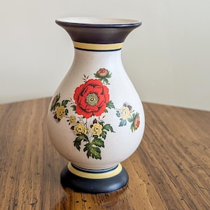 May include: A white ceramic vase with a floral design. The vase has a black rim and base with a yellow stripe. The flowers are red, yellow, and green.