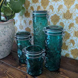 Peut inclure: Un ensemble de quatre bocaux de rangement en verre bleu sarcelle avec des motifs de fruits en relief. Chaque bocal est doté d'un couvercle en verre transparent, d'une fermeture à étrier en métal et d'un joint en caoutchouc pour l'étanchéité.