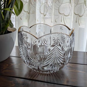 May include: A clear glass bowl with a scalloped rim and gold trim. The bowl features etched designs of trees and other patterns. The bowl is sitting on a dark wooden surface.