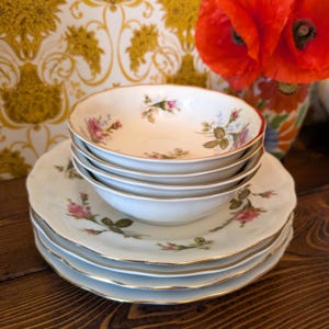 May include: A stack of white ceramic bowls and plates featuring a delicate pink rose floral pattern with green leaves and thin gold trim along the scalloped edges. The set includes multiple bowls nested inside each other, resting on a stack of plates.