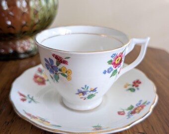 May include: White teacup and saucer set with a floral pattern in red, blue, and yellow. The cup and saucer have gold trim. The saucer has a scalloped edge. The teacup is on a wooden surface.