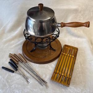 May include: A stainless steel fondue pot with a wooden handle and lid, set on a black metal stand and a wooden base. Includes a set of fondue forks and a box of forks.