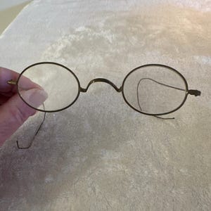 May include: Antique round eyeglasses with a thin, dark metal frame. The lenses are clear, and the frame has a bridge and temples. The glasses are held by a hand, suggesting they are being displayed or offered for sale.