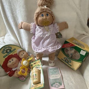 May include: A vintage Cabbage Patch Kids doll with blue eyes, wearing a lavender dress and a bonnet. Accessories include a pacifier, a bottle, a rattle, and a birth certificate. The doll is surrounded by original packaging.