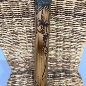 May include: A tall, cylindrical container with a black lid and base. The body is a light brown with vertical lines and a dark brown floral design. The lid is wrapped with a dark, textured material. The container is standing in front of a woven wicker chair.