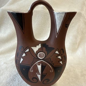 May include: Brown ceramic vase with a unique handle and decorative geometric patterns in black and white. The vase features a rounded body and a distinctive opening. The design includes stylized shapes and lines.